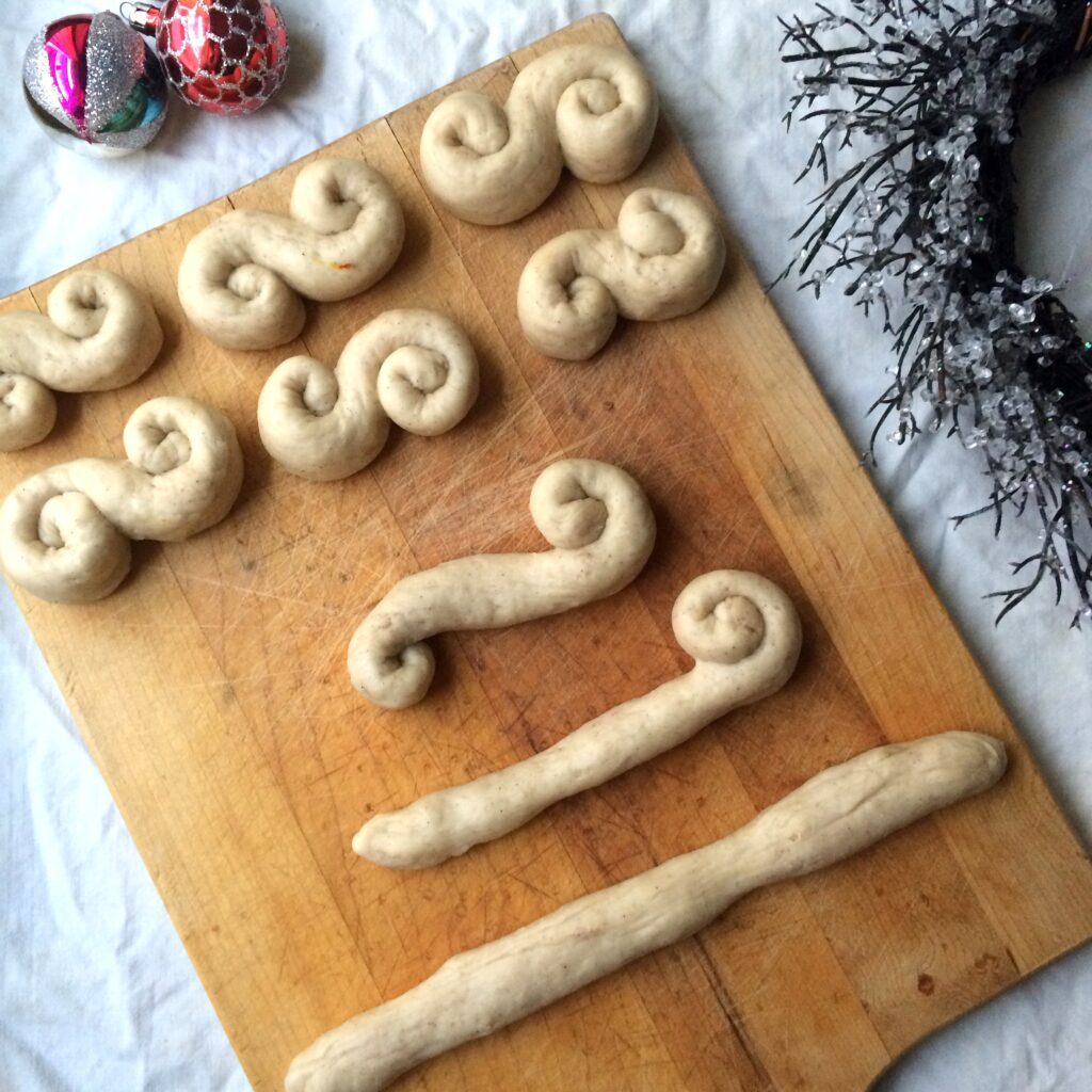 A wooden board displays several unbaked, twisted dough shapes in the form of curled "S" shapes, arranged neatly. Christmas ornaments and a decorative wreath with frosted twigs are placed nearby, suggesting a holiday baking theme.