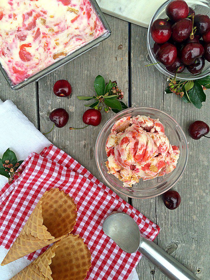 A rustic scene featuring a glass bowl of cherry ice cream, a pan with more ice cream, a bowl of fresh cherries, two empty waffle cones on a red and white checkered cloth, and a metal ice cream scoop. Scattered cherries and green leaves add to the decor on the wooden surface.