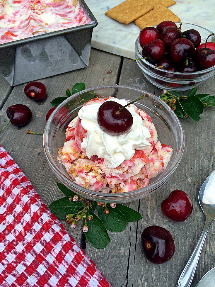 A bowl of cherry cheesecake ice cream sits on a wooden surface, topped with whipped cream and a cherry. Surrounding the bowl are fresh cherries, green leaves, a red and white checkered napkin, a spoon, and a pan of the ice cream with graham crackers nearby.