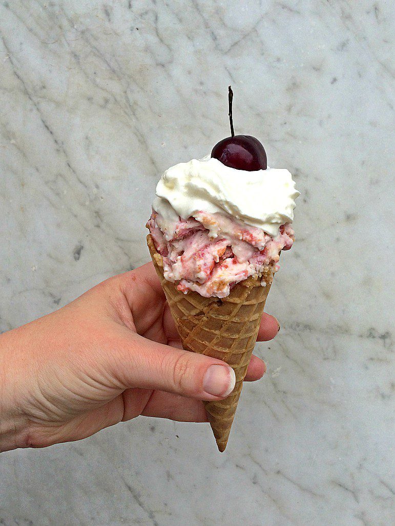 A hand holds a waffle cone filled with cherry cheeseacke ice cream topped with whipped cream and a cherry, against a light marble background.