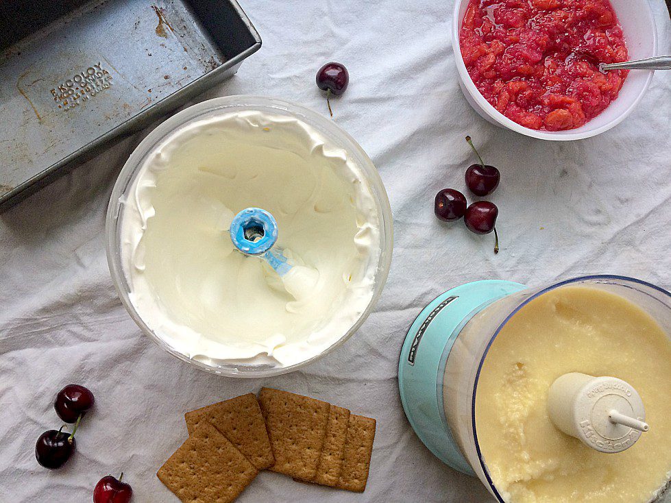 An overhead view of a dessert-making setup on a white surface. It features a whipped mixture in a food processor, a bowl of cherry mixture, several fresh cherries, a few graham crackers, and an empty baking pan.