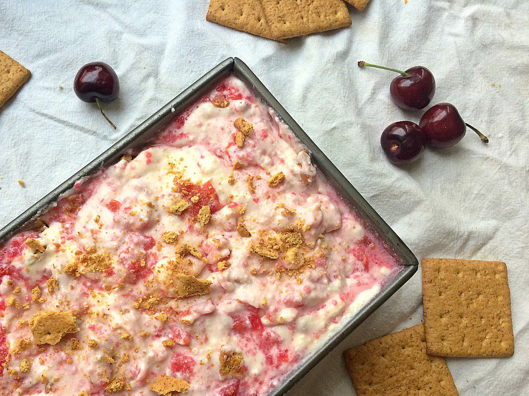 A rectangular pan filled with a creamy dessert topped with crumbled graham crackers and a pink swirl sits on a white cloth. Surrounding the pan are whole graham crackers and a few cherries, adding a pop of color to the scene.