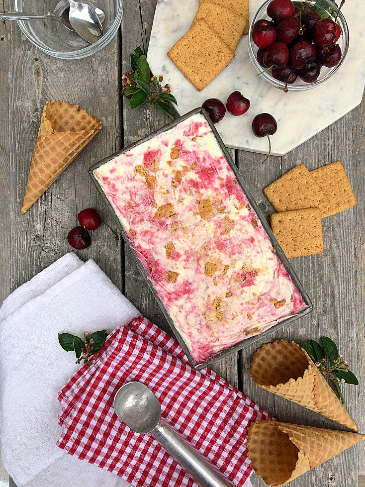 A top-down view of a dessert spread on a wooden table. The scene includes a rectangular tray of ice cream with visible layers, waffle cones, graham crackers, a bowl of cherries, and a red-and-white checkered napkin with an ice cream scoop on it.
