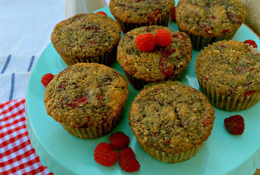 A pale green plate holds six raspberry bran muffins. Three fresh raspberries decorate the center muffin. Additional raspberries adorn the plate. The plate sits on a table with a red checkered cloth beside it.