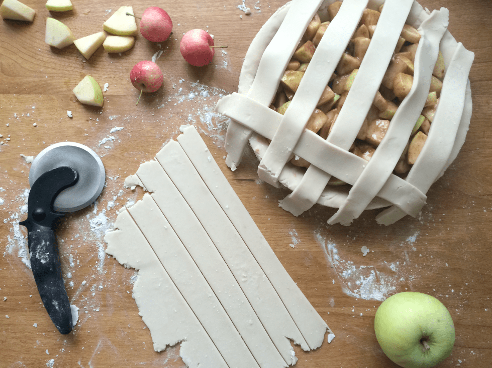 A partially assembled apple pie with lattice crust lies on a wooden table. Next to it are dough strips, a pastry cutter, small red apples, apple slices, and a whole green apple. Flour is scattered on the table.