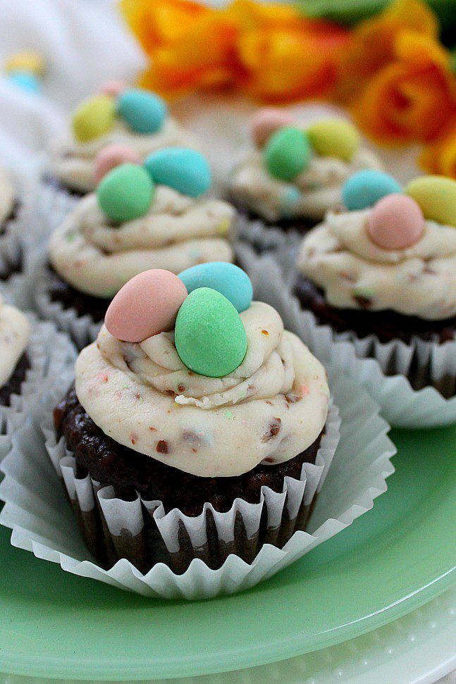 Chocolate cupcakes with swirled vanilla frosting are topped with colorful pastel candy eggs. The cupcakes are arranged on a light green plate with yellow-orange flowers blurred in the background.