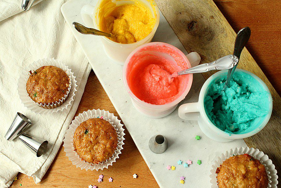 An overhead view of a baking scene with cupcakes in white paper liners, and three mugs containing yellow, red, and blue frostings with spoons inside. Metal piping tips and star-shaped sprinkles are scattered around on a wooden surface and a white cloth.