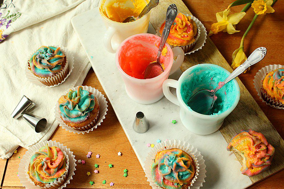 Colorful frosted cupcakes are arranged on a wooden table with three cups of frosting in yellow, red, and blue, along with utensils. Star-shaped sprinkles are scattered around, and there are a few piping tips visible. Yellow daffodils add a decorative touch.