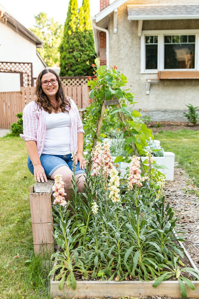 Woman with white shirt and jean shorts sitting next to a raised bed cut flower garden.