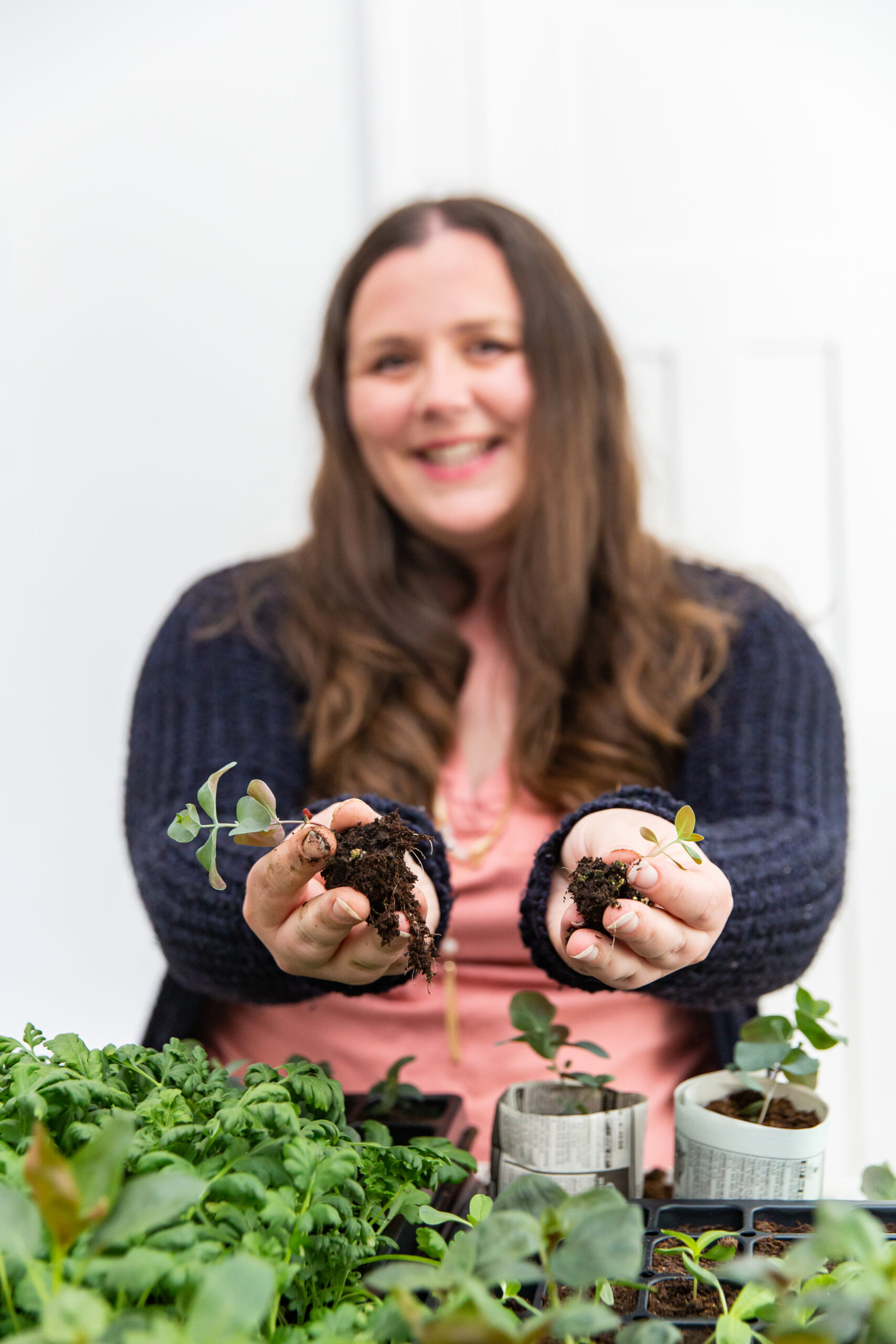 A woman with long brown hair smiles at the camera while holding small eucalyptus plants in her hands. She is standing behind a table with various plants and seedlings. She is wearing a pink top and a dark cardigan, and the background is blurred.